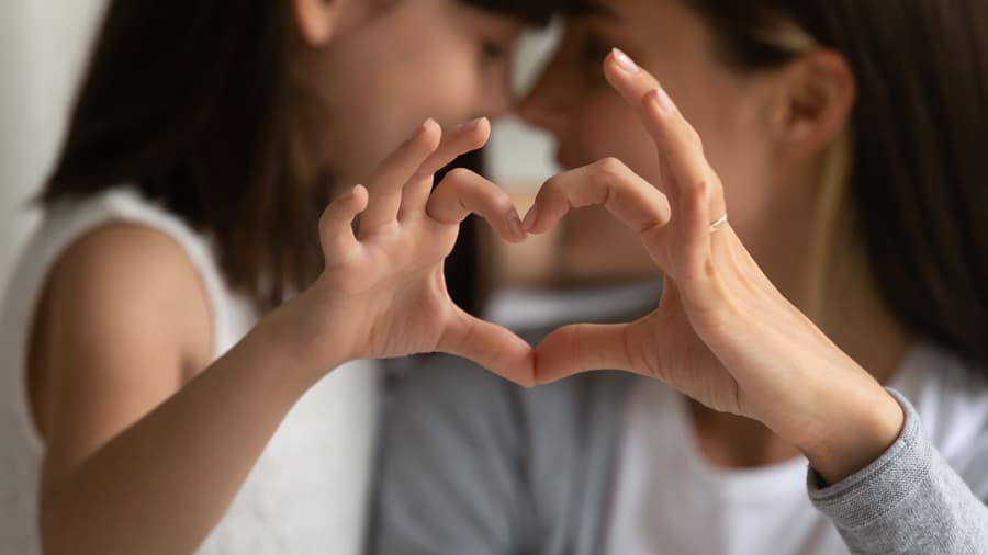 A close-up of a mother and daughter forming a heart shape with their hands, smiling and leaning their heads together in the background.