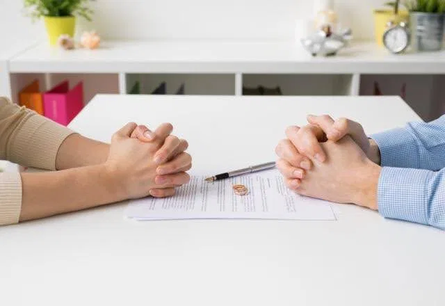 Two people with clasped hands sit across from each other at a table with divorce papers and rings between them