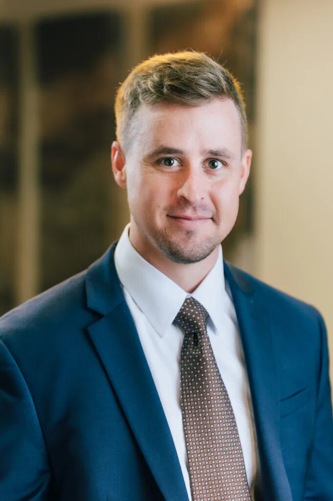 RYAN D. SARLO, in a suit and tie stands confidently in front of a plain wall, exuding professionalism and poise.