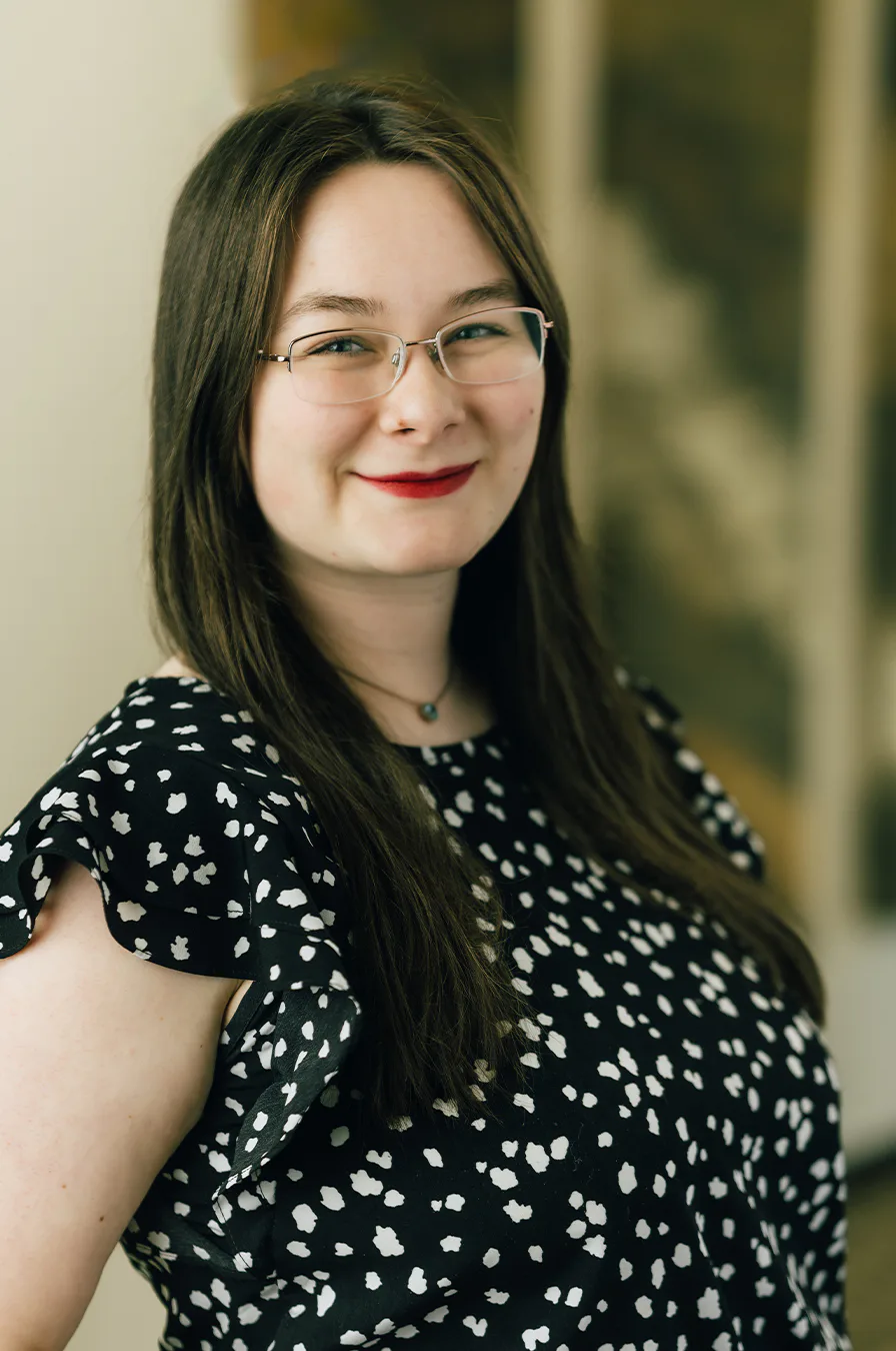 A woman wearing glasses and a polka dot dress, smiling and standing confidently.