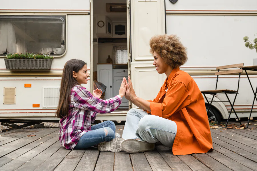 A cheerful mother and daughter sitting outdoors on a wooden deck in front of a camper van, smiling as they play a hand-clapping game.