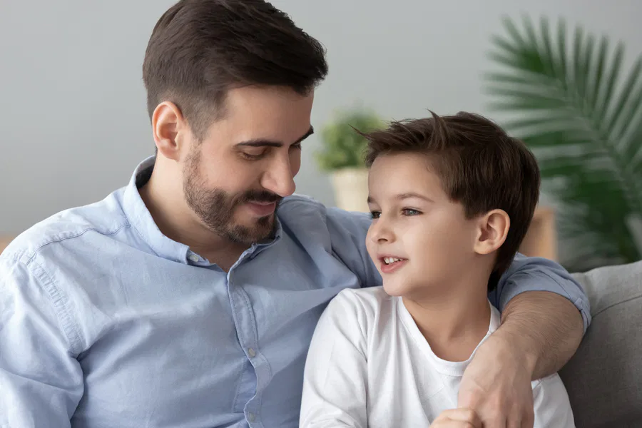 A father and son sitting on a couch at home, smiling and having a heartfelt conversation, surrounded by a cozy and green indoor setting.