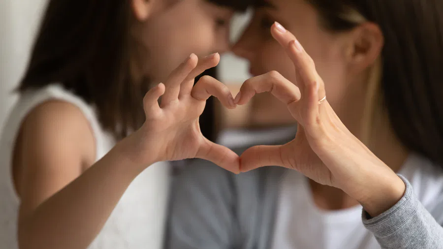 A close-up of a mother and daughter forming a heart shape with their hands, smiling and leaning their heads together in the background.