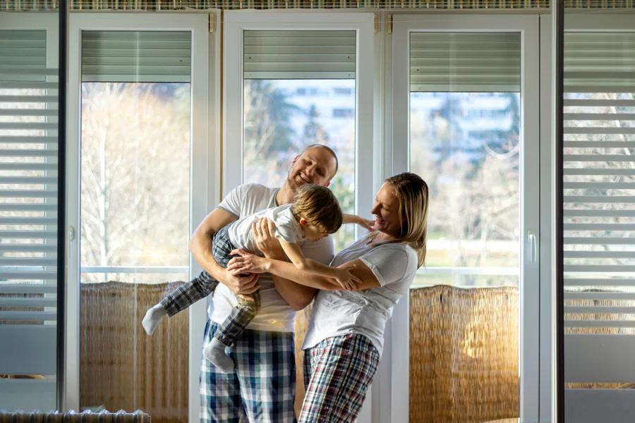 A cheerful family of three, dressed in casual pajamas, playing and bonding together in a bright living room with large windows and a sunny outdoor view.