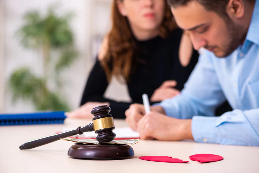 A couple signing divorce papers with a broken heart symbol and gavel on the desk, representing family law proceedings. Burton Family Attorneys.