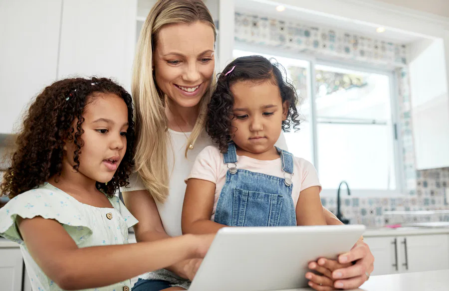 A mother happily engaging with her two young daughters while looking at a tablet in a bright, modern kitchen, representing family bonding and technology use.