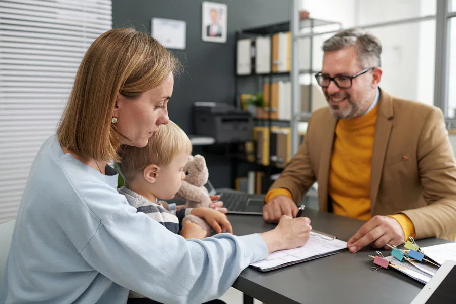A man and woman sit at a desk, engaged in conversation, while a child plays nearby, creating a warm family atmosphere.
