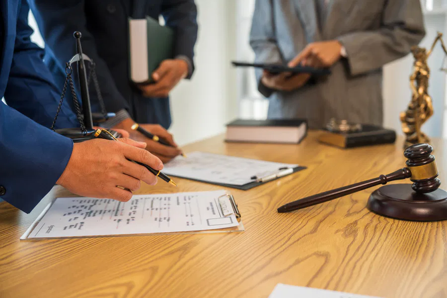 Legal professionals discussing documents on a wooden desk, featuring a clipboard, a gavel, scales of justice, and law books, symbolizing a legal consultation or courtroom preparation.