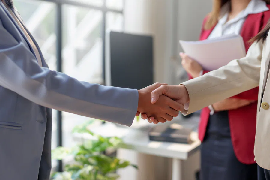 Close-up of two individuals in business attire shaking hands in a professional office setting, with a third person holding documents in the background.