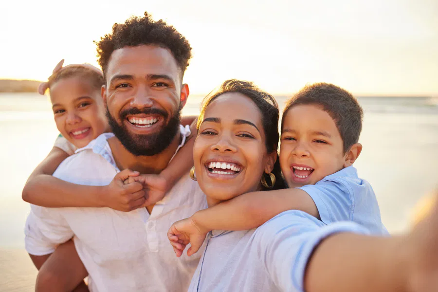 A happy family of four poses for a selfie on the beach at sunset, with the parents smiling and holding their two young children on their backs.