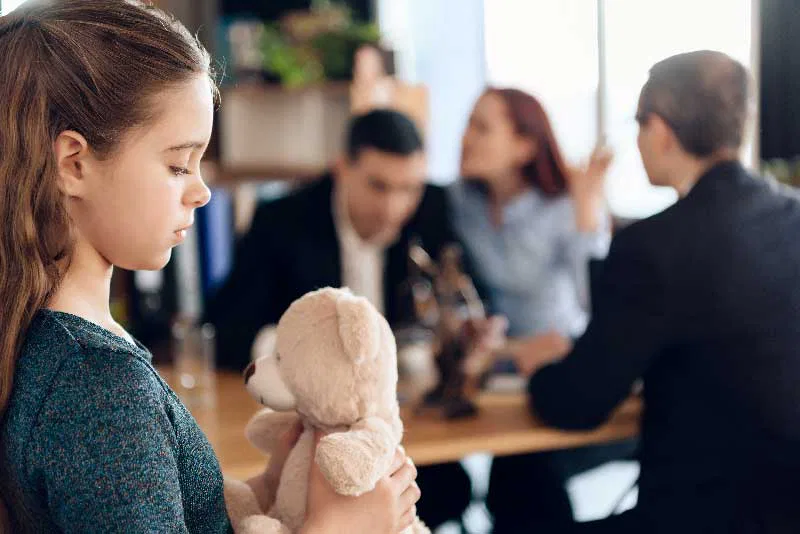 A young girl with a teddy bear stands in front of a crowd, showcasing innocence and warmth amidst the gathering.