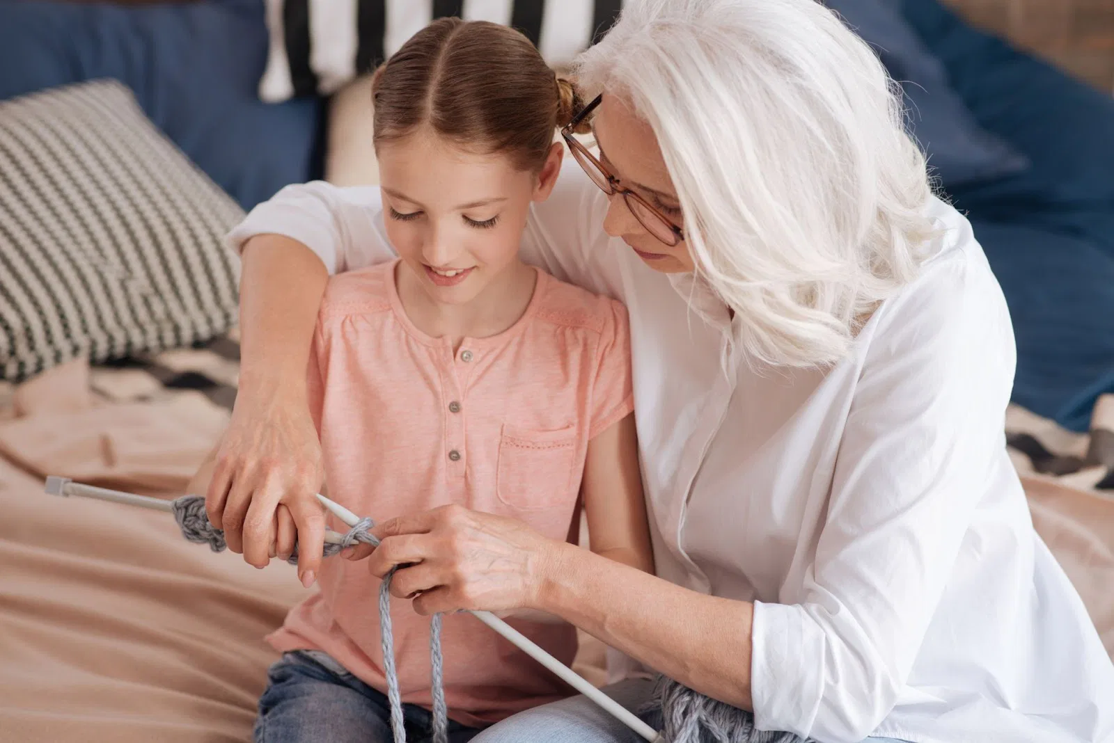 A child in a pink shirt learns to knit with an adult in a white shirt on a bed with striped pillows and a blue couch in the background.