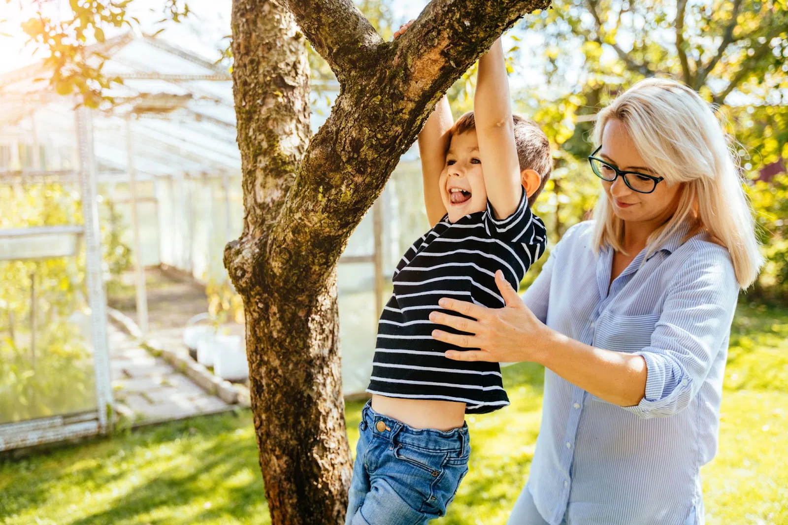 A child in a striped shirt climbs a tree while an adult in a striped shirt assists, with a greenhouse and grassy area in the background.