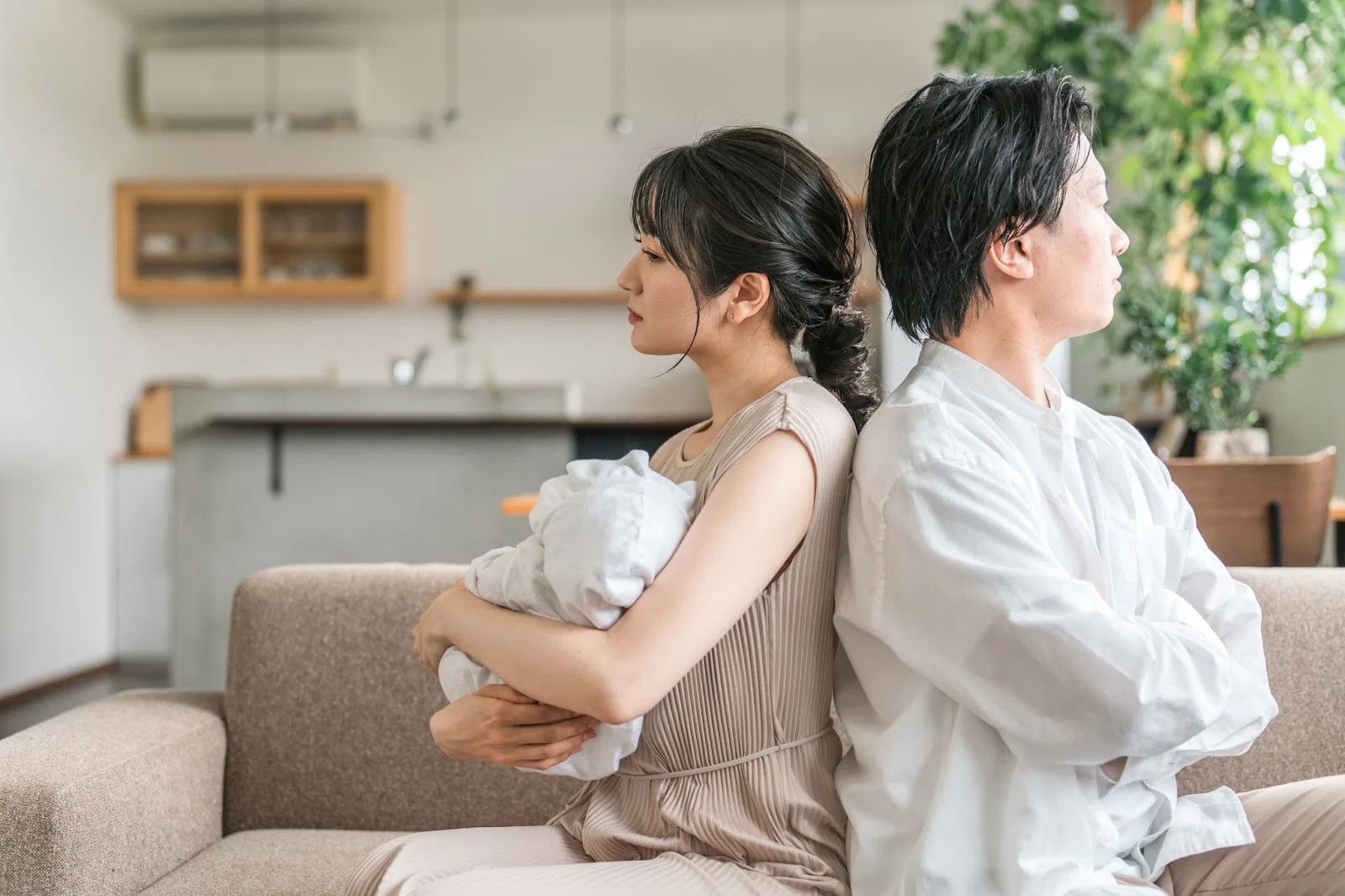 Two people sit back-to-back on a couch, one holding a baby, in a modern living room.